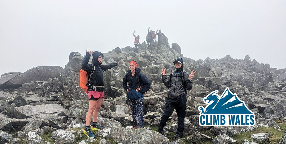 A group ladies with two fingers in the air, atop the rocky summit of Carnedd Gwenllian (Peak 2 for this group).