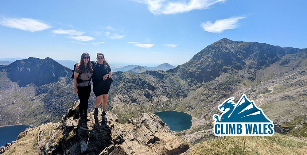 Two young ladies in hiking gear stood atop a pinnacle with Snowdon summit and Lliwedd in the background