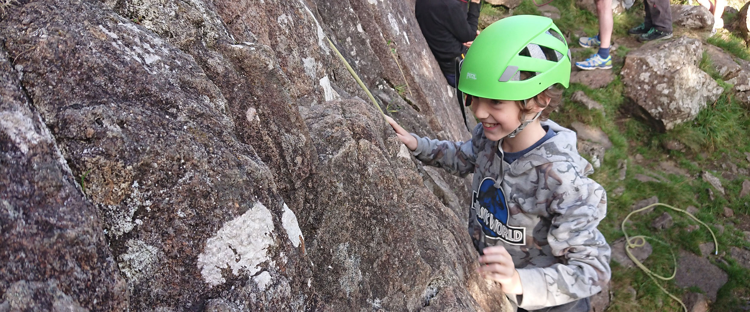A young boy at the foot off a cliff, wearing a climbing helmet, and a beaming smile
