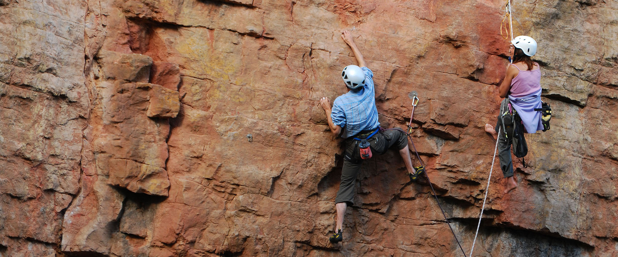 A climbing instructor hangs from a rope and offers advice to their client. The client appears to climbing through a difficult section on a sheer face